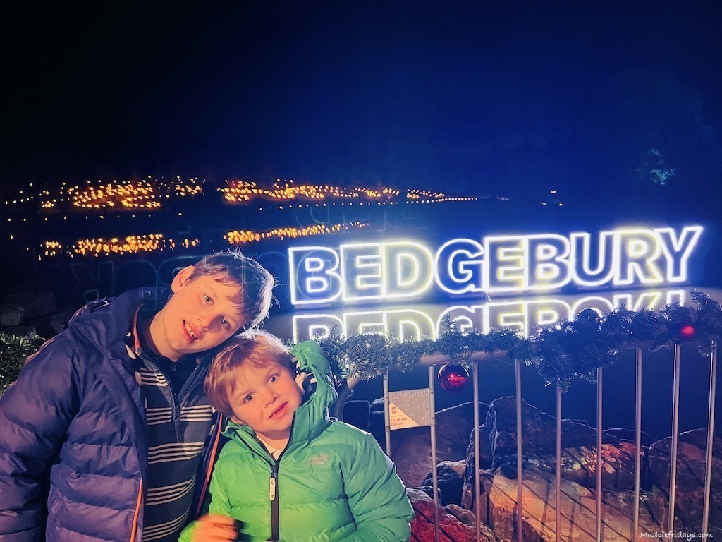 The boys standing in front of the Lake at Bedgebury Pintetum in Kent with the lit up Bedgebury sign for the Christmas light trail