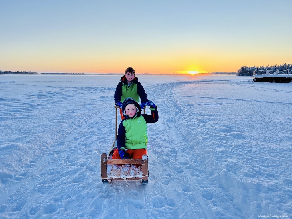 The boys sitting on a sledge in the middle of a frozen lake with the sun setting in the background