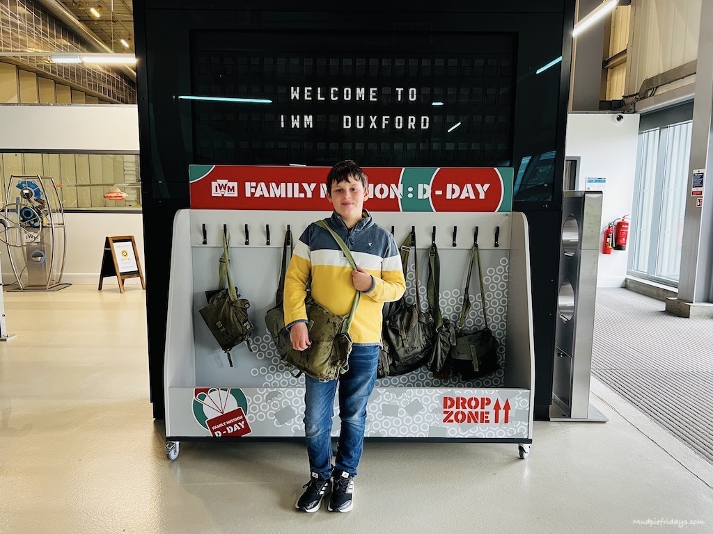 Charlie in front of the welcome to IWM Duxford sign in hangar 1 with the D day activity pack