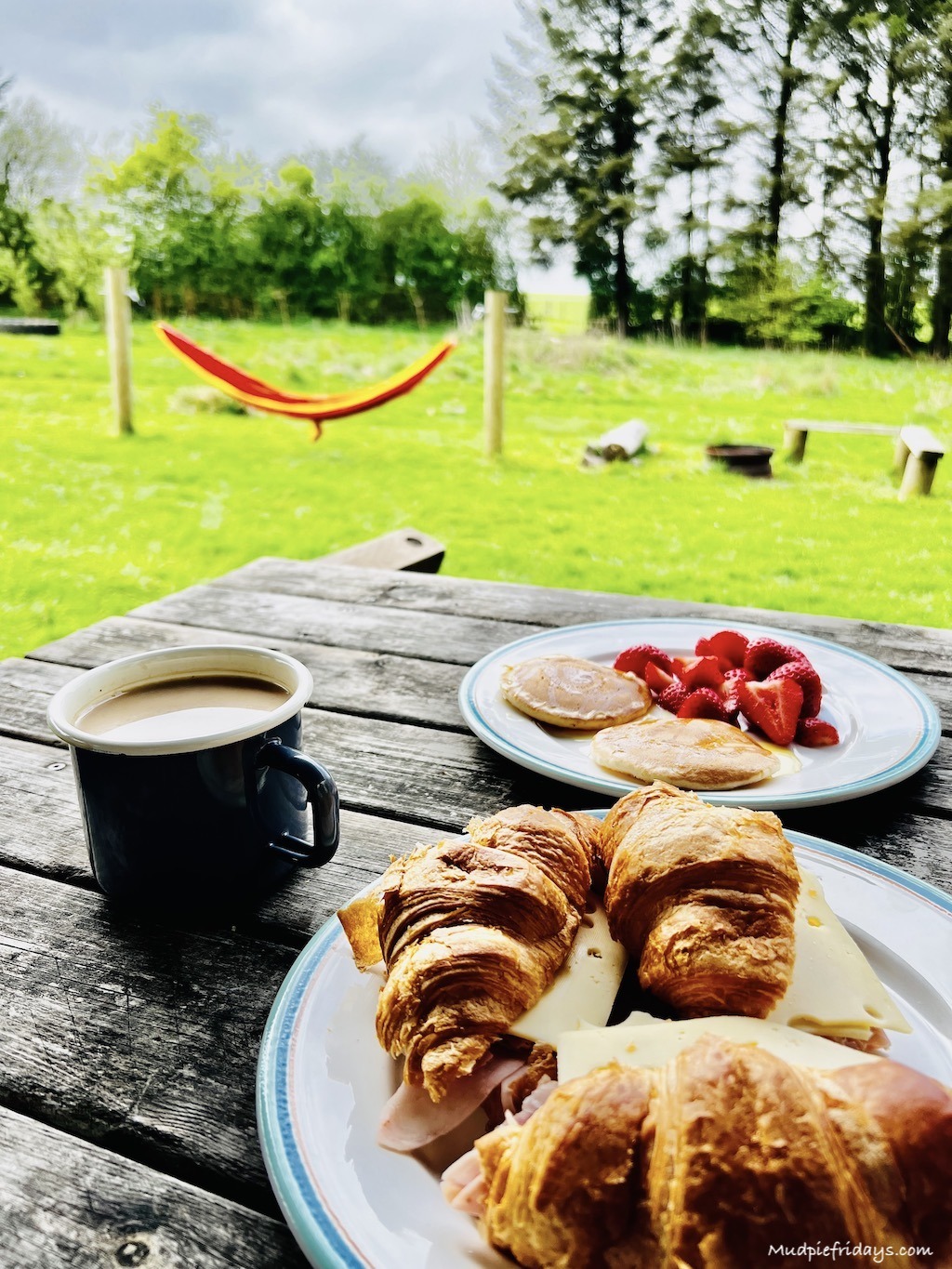 Breakfast with a view - camping mug with pancakes and strawberries and croissants with a hammock and fire pit in the background